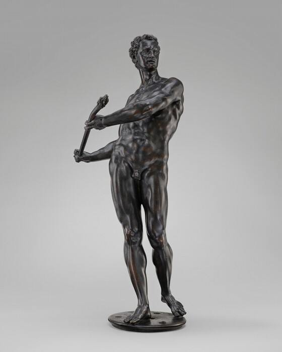 The sculpture depicts a standing male figure posing dynamically with his torso slightly twisted and his head turned to the side. The figure holds an object resembling a club or staff in both hands, crossing his body at an angle. The figure is nude with detailed musculature and a well-defined physique. The sculpture is made of bronze with a dark, metallic color, subtle variations in shading, and a reflective surface. The sculpture stands on a round, tiered base.