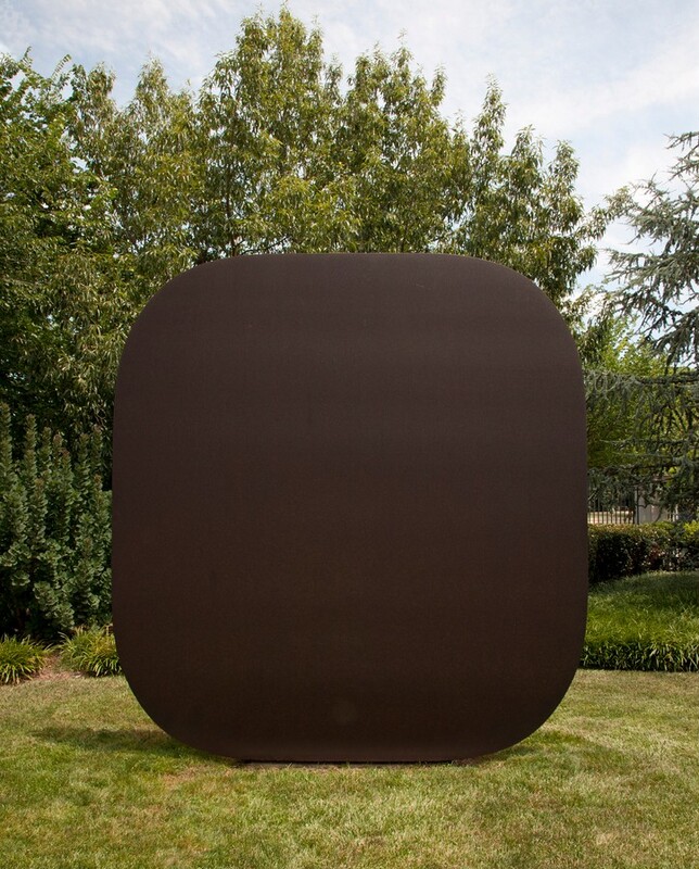 A steel square with rounded corners sits upright on a grassy lawn in front of a row of trees under a blue sky. The metal is weathered and lightly pitted to a dark, bronze brown. It looms over the bushes behind it.