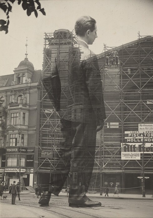 This is a photograph of a street scene with an overlay of a large figure. The image displays a city street view combined with the semi-transparent image of a well-dressed individual. In the background, there is a building under construction surrounded by scaffolding, along with advertisements and signs in German. The superimposed figure is a play on scale and transparency, suggesting a surreal quality to the photograph. The artist used a double exposure technique to blend these elements, creating a unique visual narrative.