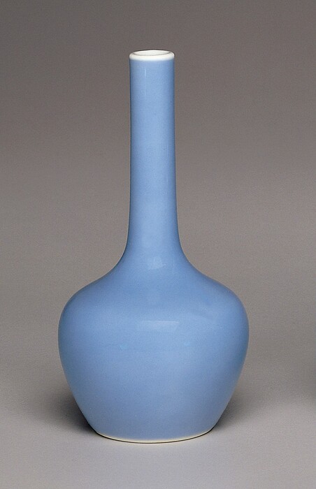 The image shows a tall, rounded vase with a narrow neck as the largest and central object. The vase is predominantly a soft pastel blue color with a slight sheen on its surface, while the background is a neutral gray. Positioned directly in the center of the frame, the vase appears upright with its base slightly wider than its tapering neck that extends upward.