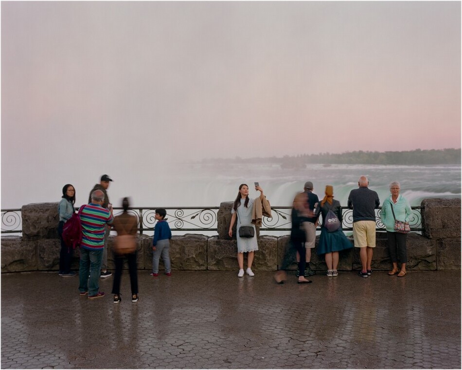 The image shows a group of people standing on a paved surface with a stone and wrought iron railing nearby. In the background, there is a misty waterfall merging into a pink sky, with green trees in the distance. Some individuals appear blurry due to motion. The color scheme includes earthy tones, pastel shades in the sky, and vibrant greens and whites from the natural elements.