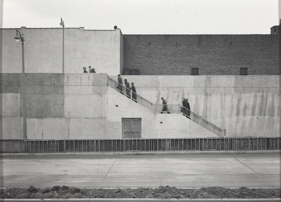 The image shows an urban stairway integrated into a building facade. The composition features a concrete structure intersecting with industrial brick walls. The staircase cuts diagonally through the scene, beginning at the lower right and rising upward to the left. The concrete wall is divided into sections of different shades, likely from weathering. A large lamp post is on the left, and a street with a well-paved road is visible in the foreground. A small patch of vegetation can be seen at the bottom, with a pale gray sky overhead creating a monochromatic tone.