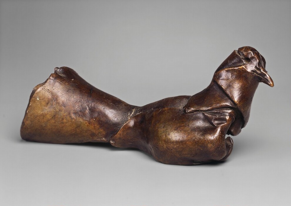 The sculpture depicts a bird in a relaxed pose with its wings sculpted closely along its body. The piece is made of a smooth bronze material, giving it a rich brown hue with natural patina. The bird's form is abstract with simplified features, creating an elegant and organic look. The focus is on the overall shape rather than precise details. The sculpture is small, potentially tabletop-sized, engaging with viewers at eye level.