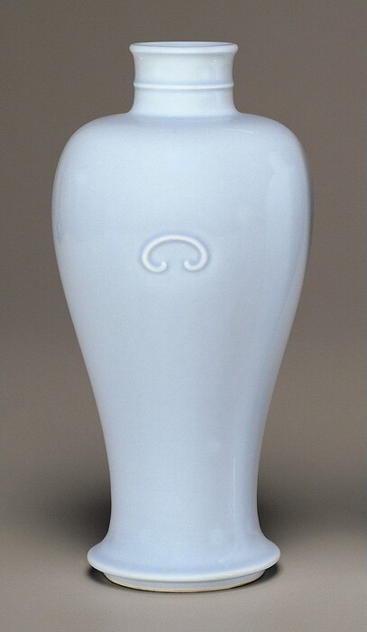 The sculpture is a vase with a simple design. It is white and appears to be made of porcelain or ceramic material with a glossy finish. The vase has a rounded, tapered body that extends into a narrower neck and flares out slightly at the mouth and base. An embossed subtle decorative element can be seen near the upper portion of the vase. The overall appearance of the vase is smooth and symmetrical, reflecting light evenly across its surface.