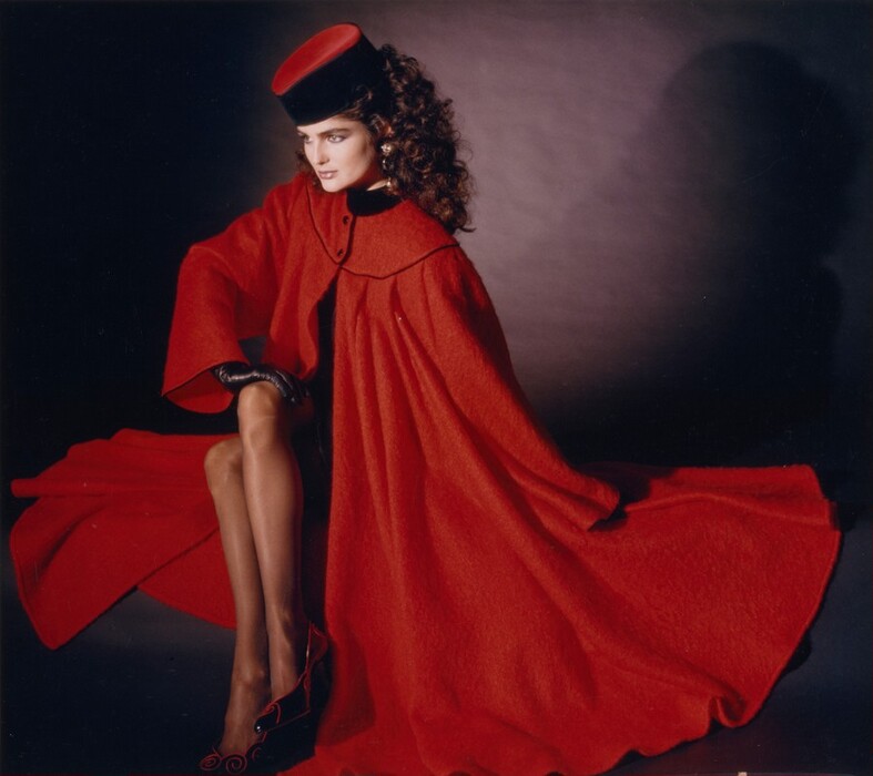 This photograph shows a person seated with their legs crossed. The person has pale skin and curly, voluminous dark hair around their face. They are wearing a long, flowing red coat and a tall red hat. They are also wearing black gloves, red shoes, and large earrings. The background is a dark gradient.