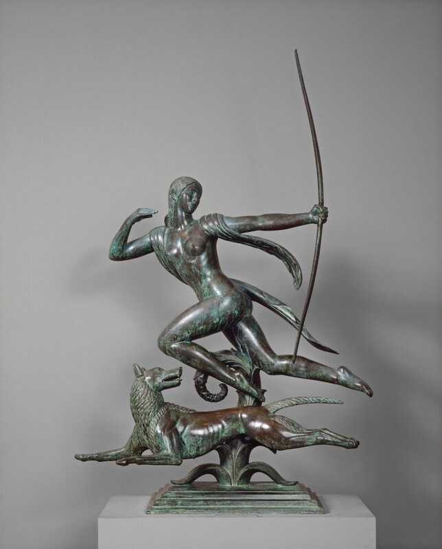 A woman holding up an archery bow runs alongside a sleek dog in this free-standing, bronze sculpture. Some areas of the surface are shiny brown and others have a mint-green patina. In this photograph, the woman and dog run to our left. The woman’s hips are squared to our left but she turns her torso to face us, her arms outstretched as she twists her head to look over her back shoulder. She looks to our right almost in profile with almond-shaped eyes under thin, arched brows. She has a straight nose, and her full lips are closed. Her hair falls in stylized waves close to her head, and is gathered at the back of her neck. Held up at shoulder height, her right arm, to our left, is bent back at the elbow. She holds her fingers together, as if holding a missing arrow or the string of the bow. With her other arm extending to our right, she holds up a bow as tall as she is, also at shoulder height. A sash drapes over her shoulders, and the ends trail behind her. She has smooth, round breasts over the shallow indentations of her ribcage. Her hips and thighs are smooth, her left knee bent in front of her and her other leg extended behind her, in midstride with her toes pointed. The muscular dog also runs with legs outstretched to our left, and it turns its head back to our right. Sharp teeth line its parted jaws, and its tongue sticks out over its bottom lip. The neck, the back of the hind legs, and its pointed tail are covered in thick, stylized fur. A curling, plant-like shape beneath and behind the woman and dog connects them and supports their weight. The plant grows from a stepped, rectangular base, which is shown on a gray, rectangular pedestal against a gray wall.