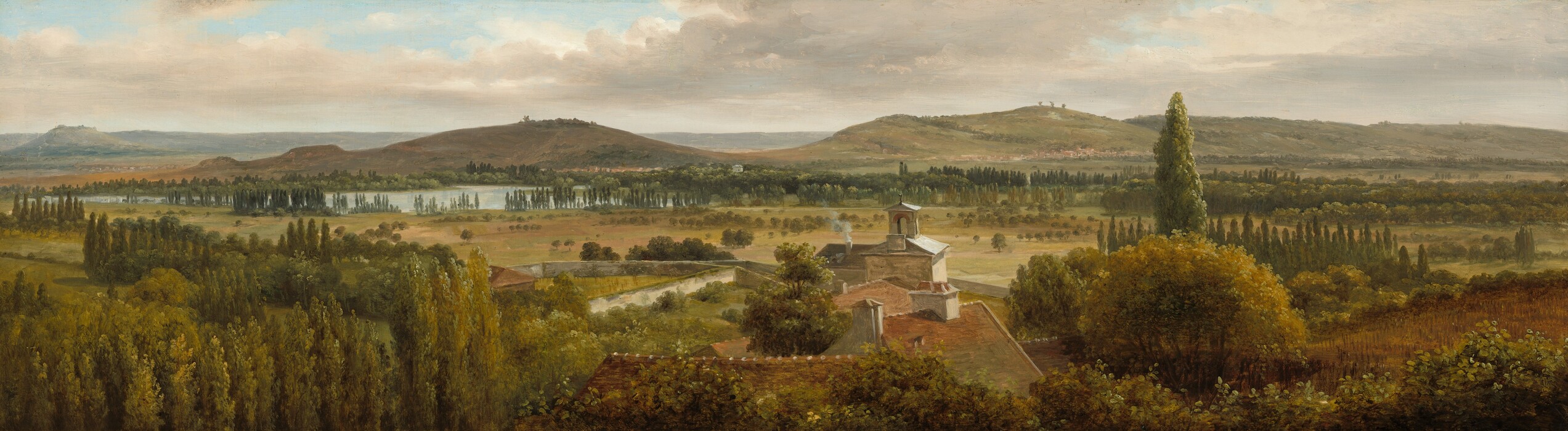 We look across the tops of trees down onto the roofs of a cluster of buildings and then a grassy expanse, a body of water, and rolling hills in this notably wide landscape painting. The trees are painted with shades of pine and olive green. At the center of the composition, the roofs are muted terracotta red with white chimneys except for the farthest structure, which has a gray roof around a lantern, perhaps indicating a church. Smoke puffs out of one chimney there. Trees with round canopies dot the pale, grassy field beyond that, which leads back to rows of tall, dark green cypress trees. A ribbon of ice blue across the left third of the painting indicates a body of water before russet-brown and muted sage-green hills roll into the deep, hazy distance. The horizon comes about two-thirds of the way up the painting, and glimpses of pale blue sky are visible through breaks in a blanket of pale gray and cream-white clouds above.