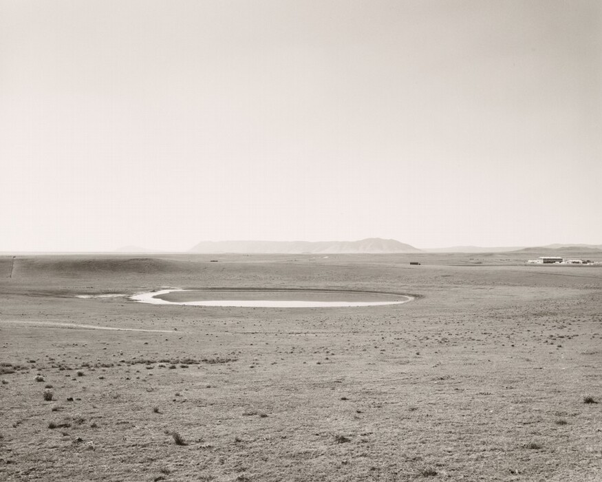 The image shows a flat landscape with sparse vegetation and tufts of grass. The horizon is high with distant hills or mountains. The style is photographic, black and white. There is a circular pond reflecting the landscape. The color palette is monochromatic gray with structures in the distance to the right. The sky above is light gray.