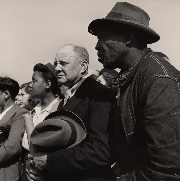 The image shows four people standing side by side. In the center is a bald man holding a rolled document and a hat. To the left is a woman partly obscured, and to the right is an older man wearing a hat and a jacket. Faint outlines of other individuals are visible in the background. The sky is clear and bright.
