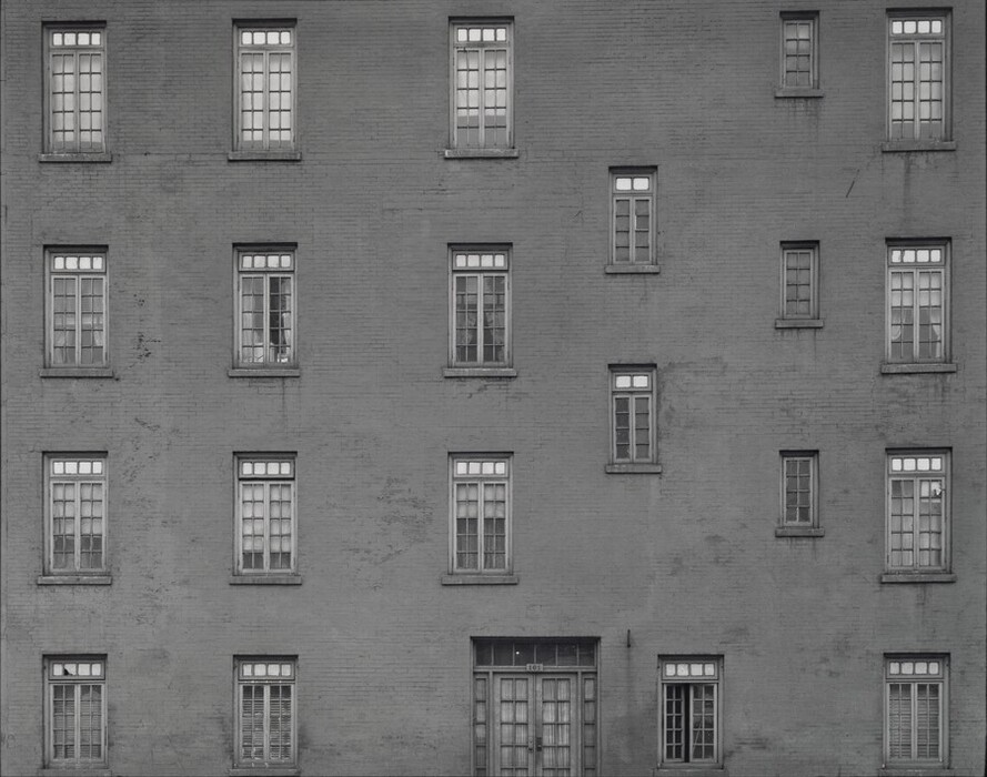 This is a photograph of a building with a brick facade and multiple windows. The image shows a symmetrical arrangement of windows on a plain brick wall. The windows are rectangular with white frames, organized in four rows with uniform spacing. The bricks are detailed, creating a textured surface. The composition emphasizes the geometric and structural elements of the architecture.
