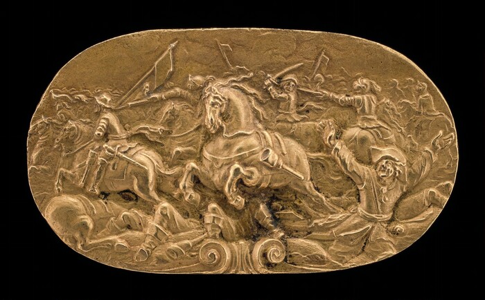 The sculpture shows a battle scene with cavalry soldiers engaged in combat. The soldiers and horses are intricately carved with details such as flowing manes and muscular builds, depicting movement and action. The soldiers are dressed in historical attire and wielding weapons. The sculpture is made of metallic material with a golden hue, creating depth and highlighting fine details. The scene is carved in relief against an oval-shaped golden background.