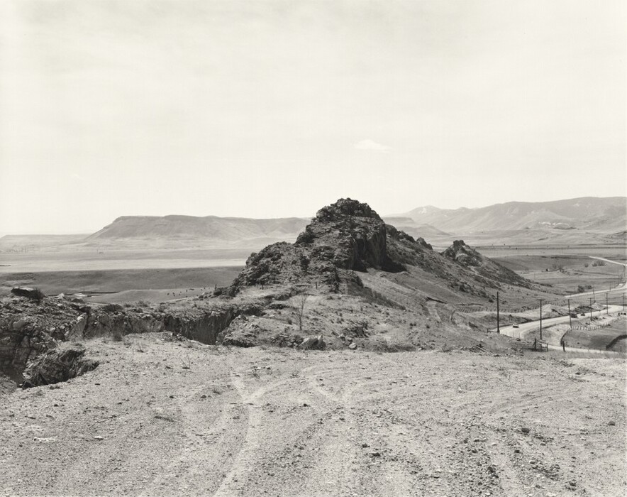 The image is of a rugged, arid landscape with an elevated vantage point. The horizon line is high, with distant mountains barely interrupting the flat sky. The terrain is rocky and sparse, with a distinctive outcrop of stone to the right. A winding road runs near the rock formation, curving through the landscape. The palette is monochromatic. In the distance, plains stretch out towards hills and mountains, giving depth to the scenery.