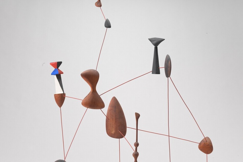 This sculpture is made up of ten shapes connected with rigid, straight brown wire to create an interlocking, web-like formation that rests on the floor. Five of the shapes appear to be raw wood and the other five are painted. Four of the painted shapes are dark gray. The fifth painted piece, at the upper left in this photograph, has a wooden cone-shaped tip pointing downward beneath a black and white mid-section, and an angular, hourglass-shaped tail painted in triangular blocks of red, blue, and black. The other wooden and painted shapes range in size and form, with an almond-shaped piece at the center, rounded pieces throughout, and two rounded hour-glass shaped pieces near the upper left.