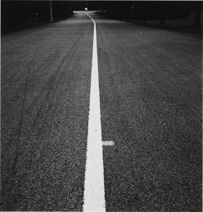 The image shows a straight-on view of an empty wide street. The horizon is in the distance where the road gently curves and disappears. The image is a photograph capturing the surface texture of the road with crisp clarity. The color palette is grayscale, contrasting the dark asphalt with the white line stretching along the road's length. The focus is on the symmetrical alignment and the interplay of light and shadow.
