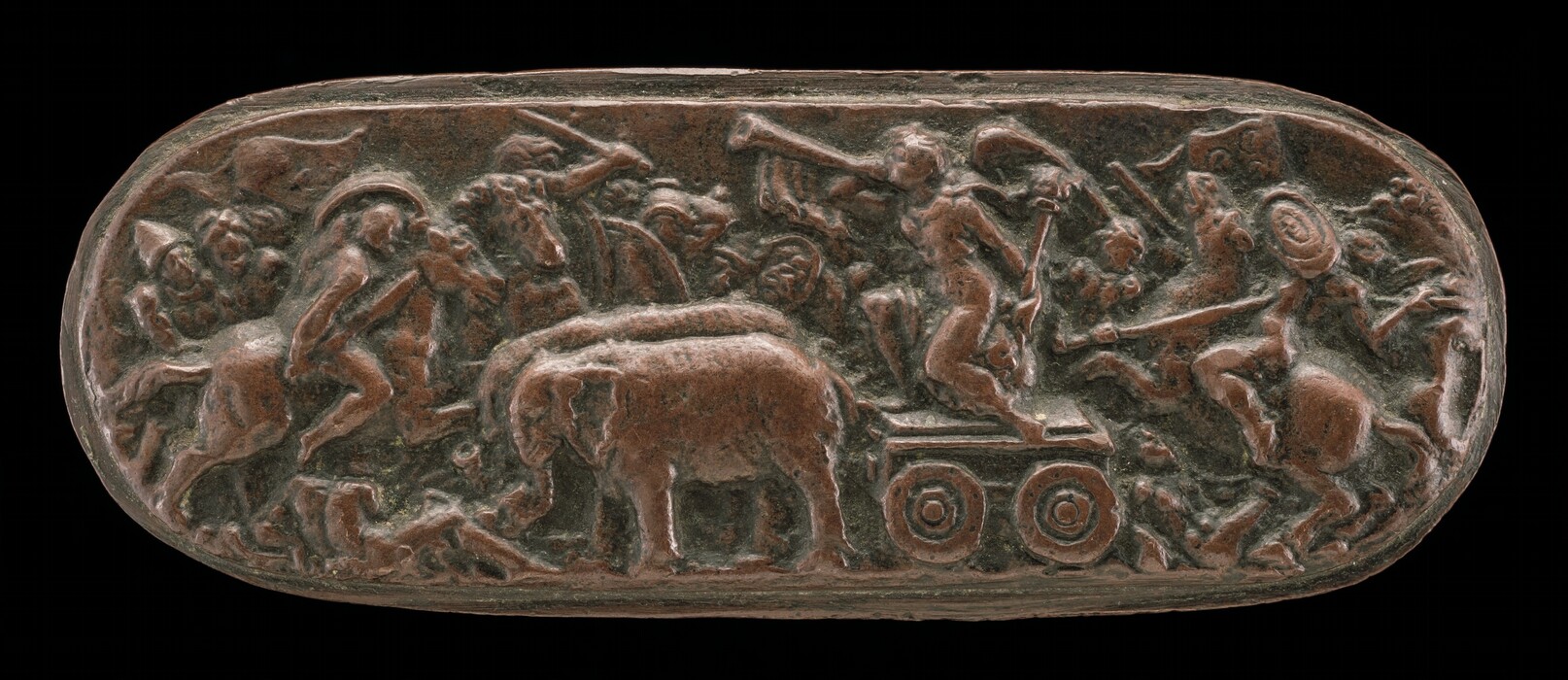 The sculpture depicts numerous human and animal figures in dynamic poses. There is an elephant on a wheeled platform in the center. The figures are engaged in various actions such as riding horses or wielding weapons like swords and shields. The sculpture is crafted from a reddish-brown material, likely bronze or a similar metal, with a dark patina. It is in low relief on an oval-shaped background, suggesting a narrative captured in miniature form. The detailing conveys movement and action to the piece.