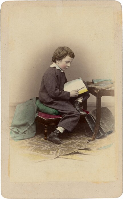 The image shows a young boy seated on a cushioned stool, reading a book. He has short, curly brown hair and is wearing a dark outfit with a white collar. Beside him lies an overcoat or a piece of cloth on the stool. Nearby is a stack of blue books on a table. The background features a plain wall and a patterned rug on the floor.