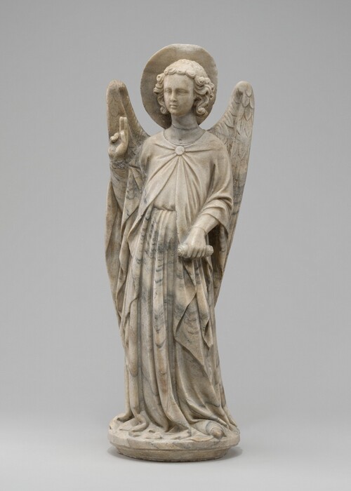 A freestanding, winged angel with a halo is carved from white marble streaked with gray. In this view, the angel’s body faces us, and the head turns slightly to our left. Curls frame a round face with delicate features in front of a flat, plate-like halo. The angel’s long robe is pinned at the chest, tied around the waist, and falls in thick folds into puddles around the feet. The angel holds one hand up with the first two fingers raised and holds a scroll down by the hip with the other hand. One pointed shoe peeks from under the robe, which almost covers the round base on which the angel stands. The sculpture is photographed against a pale gray background.