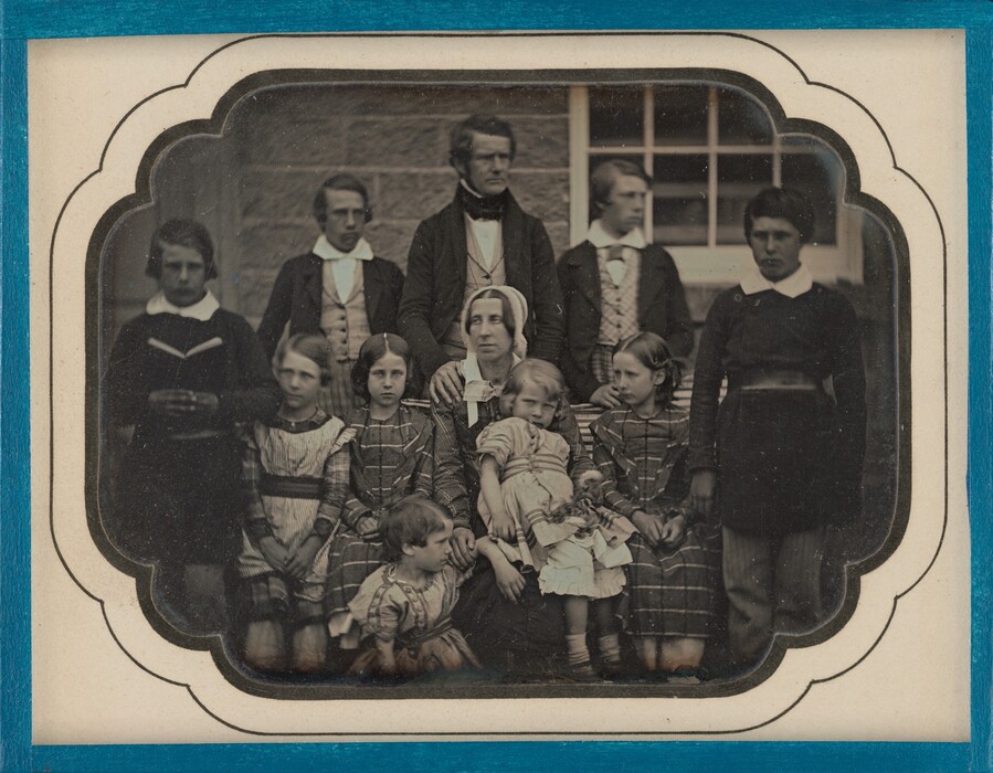 The image depicts a group portrait of eleven individuals. There are eight individuals standing at the back and three people seated in the front. At the center is a woman with a child on her lap and two more children around her. The adults are positioned around them in the back. The man standing behind the woman is dressed formally in a dark jacket, white shirt, and a bow tie. The woman wears a bonnet, and each child wears long-sleeved dresses. One of the standing men holds a small book. The boys and men wear dark suits with white collars. The background shows a stone wall and a window. The image is framed with a decorative blue border.