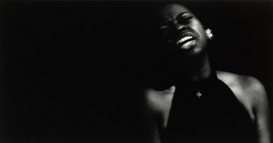 The image features a person with their upper body visible and slightly turned to the side. Their face is tilted upwards, and their mouth is open wide, displaying their teeth. The person's facial features are partially obscured due to low light. Their hair is short, and they are wearing earrings. They are dressed in a dark halter-top that reveals their shoulders, and they have a pendant necklace. The rest of the image is dark with little visible detail.
