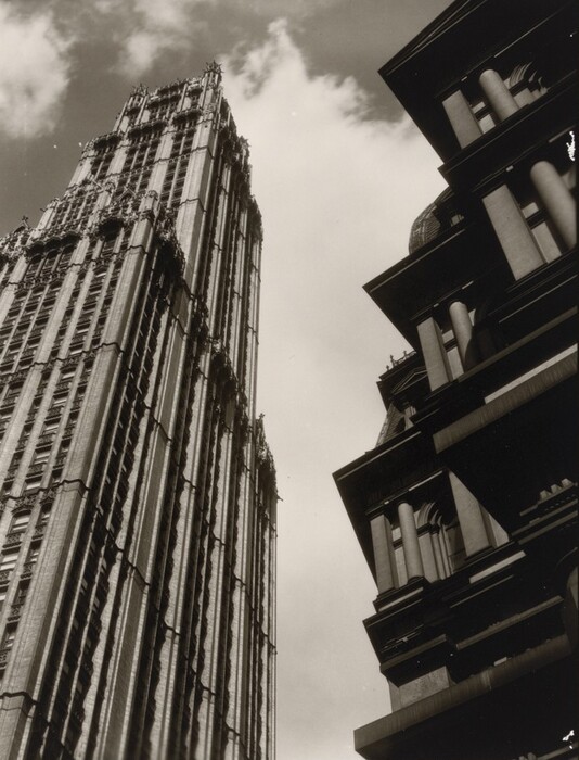 The image shows two tall buildings dominating the scene. The perspective is from a low angle, looking up at the buildings against the sky. The building on the left has intricate Gothic architectural details, while the one on the right appears stately with strong horizontal elements. The photograph is in black and white, with a contrast between the light sky and dark building facades. Clouds in the sky add a dynamic element to the composition.