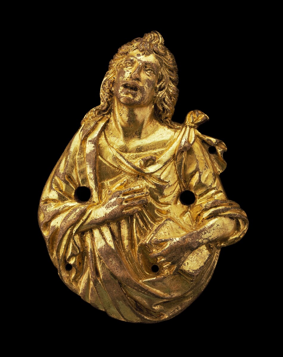 The sculpture depicts a bust of a person with long, curly hair and an expressive face looking upwards. The material of the sculpture appears to be gilded, giving it a bright golden color. The figure is draped in flowing robes with detailed folds. There are two large holes in the chest area. The figure's hand is placed across the chest. The artwork stands against a plain black background.