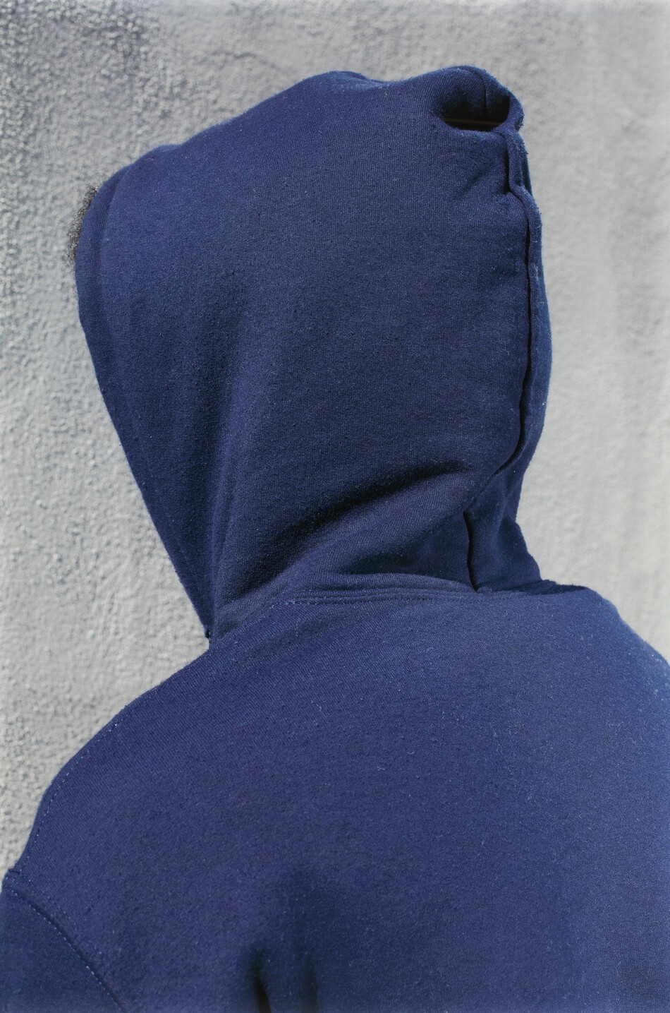 The image shows the upper body of a person from behind, with the head and shoulders visible. The person is slightly turned, facing away from the camera. Their face is not visible as the hood of their clothing is up, covering their head entirely. The person is wearing a dark blue hooded sweatshirt. The background consists of a textured gray wall. The image provides minimal context due to the obscured view of the person's facial features and details.