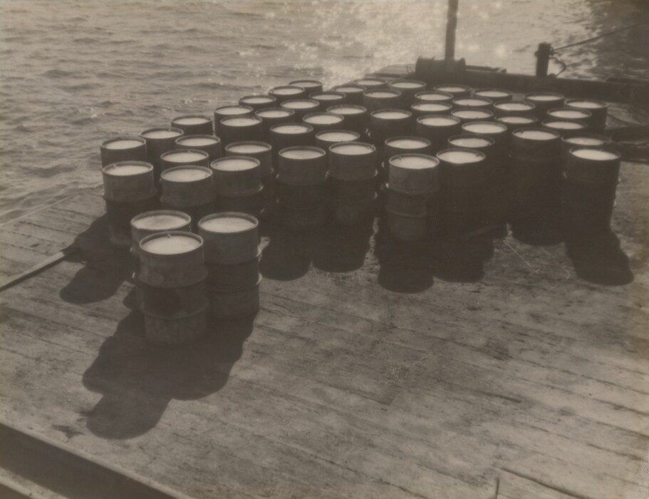 The image shows a collection of barrels stacked on a wooden dock or platform. The color palette is monochromatic, ranging from light grays to dark blacks, indicating it is a black and white photograph. The barrels are placed towards the right side of the image, with shadows cast on the platform on the left. Water is visible in the upper portion of the image, with a lightly textured surface and reflections from the light source.