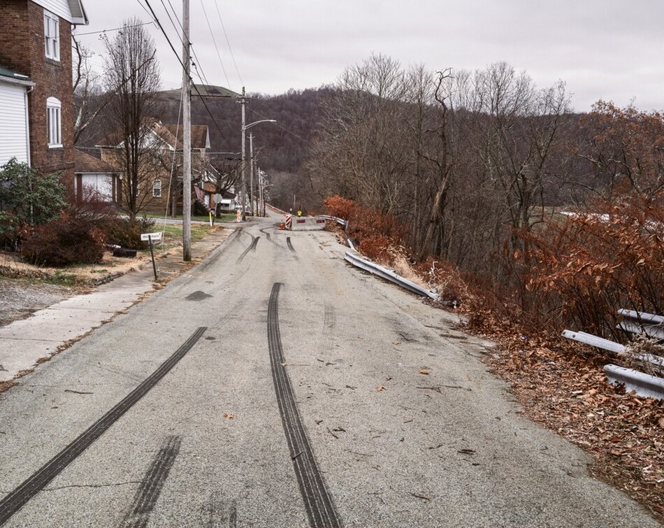 The image shows a quiet road curving gently to the right. There are houses with brick and white siding on the left, along with bare trees and shrubs. On the right, there is a metal guardrail next to a slope leading to a wooded area with leafless trees. In the distance, the road rises slightly, with rolling hills and more leafless trees visible on the horizon. The sky is overcast, casting a muted light over the scene, creating a cool color palette of grays, browns, and subtle hints of autumnal orange and red in the shrubbery.