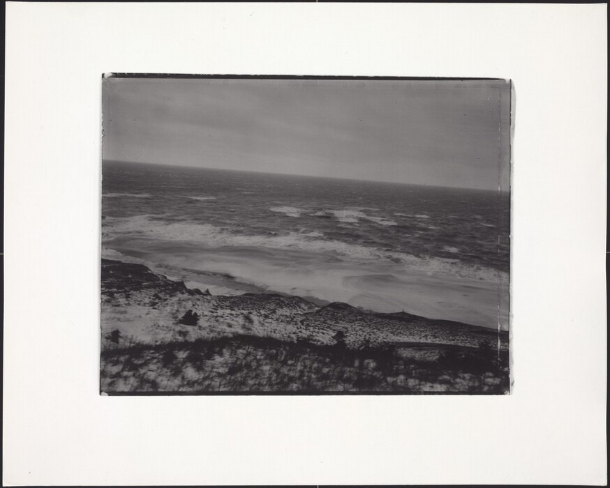 The image shows a coastal scene in a black and white photograph. The viewpoint is slightly elevated, looking out over a rugged shoreline with frothy waves crashing against the beach. The horizon line is high in the composition, where the ocean meets the sky. The photograph emphasizes contrasts and textures, highlighting the movement of the waves and the terrain of the land. In the background, the sea and sky merge into similar shades of gray, enhancing the monochromatic color palette and visual impact of the scene.
