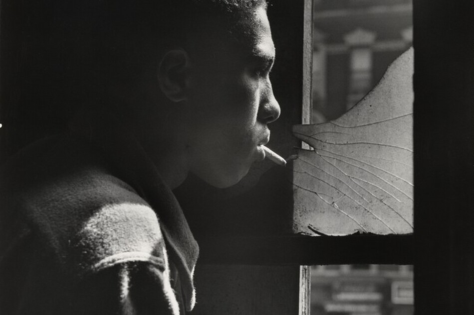A young Black man stands in profile looking out a tall window to our right in this vertical black and white photograph. The image is cropped so his head, shoulder, and upper arm fill the left half of the composition. He has short hair and wears a shirt or jacket that diffuses the light to suggest that it could be flannel or another soft fabric. With a cigarette dangling loosely from his lips, he stares through the shattered upper pane of the window and he holds his right hand across his chest. The strong light source from the right accentuates his nose, cheeks, hair, and shoulder, and the space behind him is lost in shadow. A blurred, three-story building across the street has a dark façade with white stone lintels above the windows and a mansard roof with curved dormers.