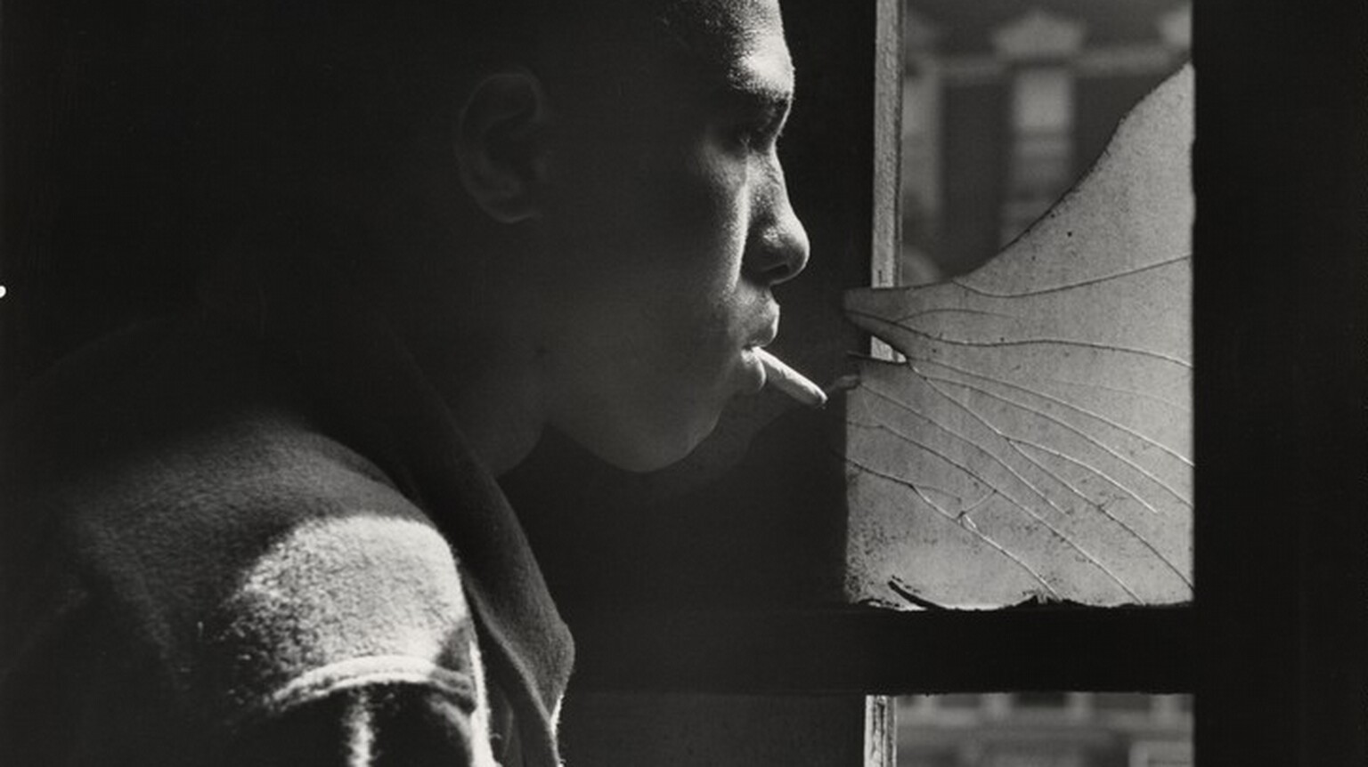 A young Black man stands in profile looking out a tall window to our right in this vertical black and white photograph. The image is cropped so his head, shoulder, and upper arm fill the left half of the composition. He has short hair and wears a shirt or jacket that diffuses the light to suggest that it could be flannel or another soft fabric. With a cigarette dangling loosely from his lips, he stares through the shattered upper pane of the window and he holds his right hand across his chest. The strong light source from the right accentuates his nose, cheeks, hair, and shoulder, and the space behind him is lost in shadow. A blurred, three-story building across the street has a dark façade with white stone lintels above the windows and a mansard roof with curved dormers.