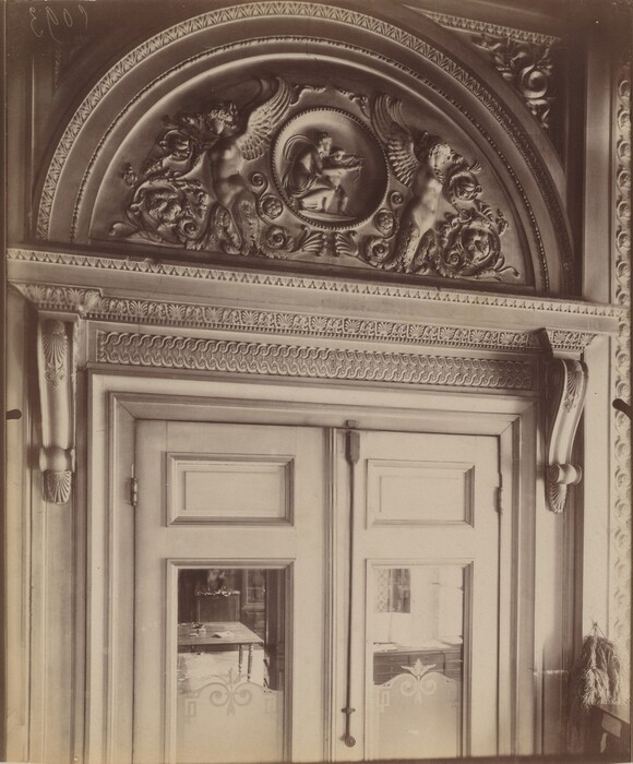 This is a photograph of an ornate doorway with elaborate architectural detailing. The image captures a richly decorated archway above a double door, featuring intricate carvings and sculptural elements. Above the door, there is a semi-circular relief with two winged figures and a central medallion surrounded by scrolling foliage and decorative patterns. The doors have glass panels with etched designs, offering a glimpse into an interior space. The surrounding framework is adorned with detailed moldings and ornamental motifs.