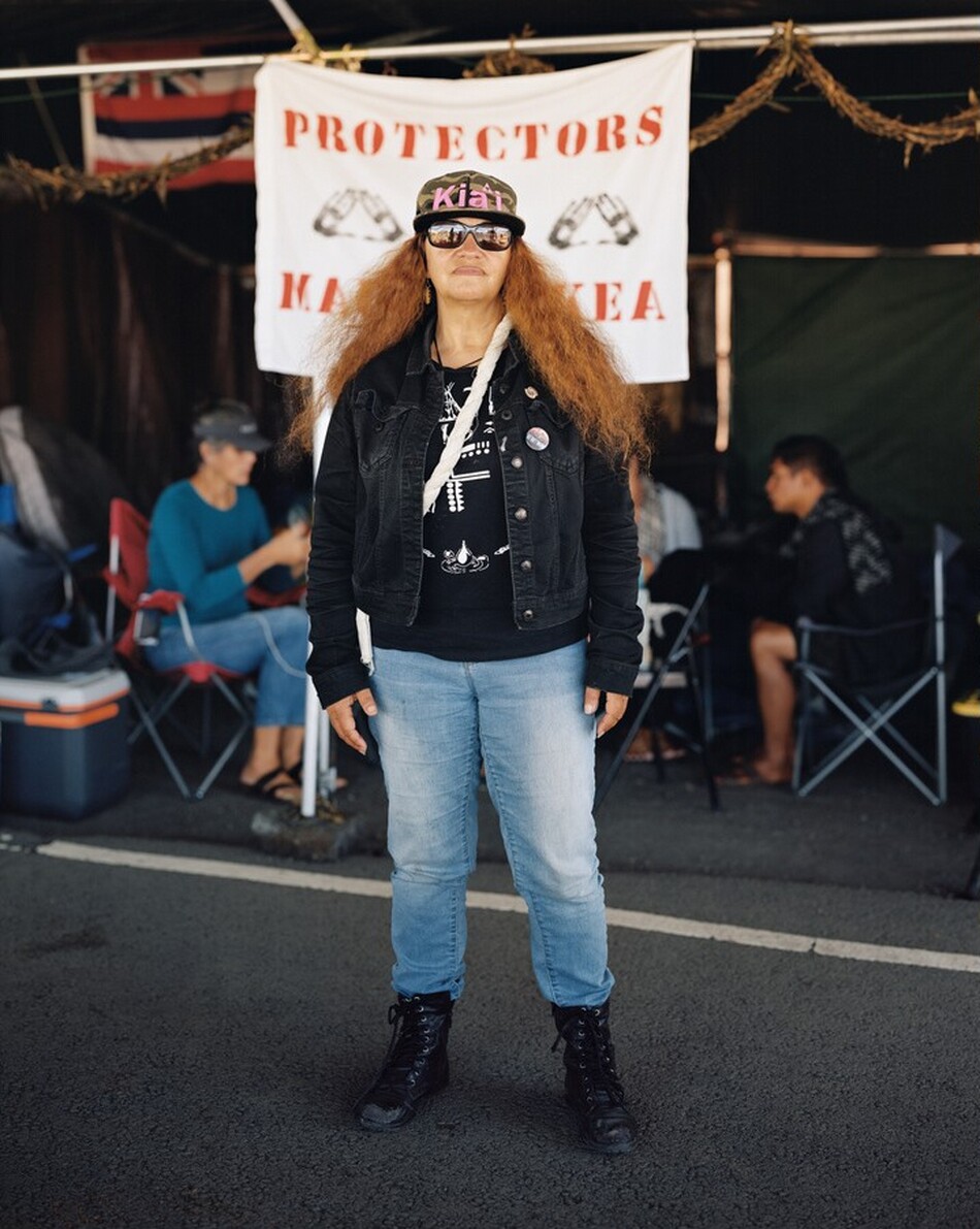 This photograph shows a person standing upright, visible from head to toe. They have lightly tanned skin and long, wavy, reddish-brown hair, and are wearing dark sunglasses. Their clothing includes a black denim jacket over a black shirt with a white design, light blue jeans, and black lace-up boots. They are wearing a cap with text on it and have a white strap across their chest and one shoulder. Circular pins are attached to their jacket, and they wear earrings. In the background, there is a banner with text and a design, including the word "PROTECTORS." In the background, there are people seated on foldable chairs with a cooler visible on the left. The scene is outdoors, with a paved surface underfoot.