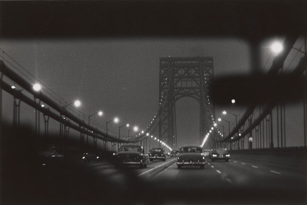 The image shows a view from inside a car traveling on a bridge with several cars heading in the same direction. The bridge is suspended between two metal structures, with streetlights along its length creating a line of glowing orbs. The steel framework of the bridge stretches into the distance, and the horizon is obscured by its architectural elements. The scene is captured in black and white, emphasizing the bridge's silhouette against a faintly illuminated sky. Smooth, fluid lines depict the bridge and vehicles, creating a strong graphic contrast in shades of grey and black.