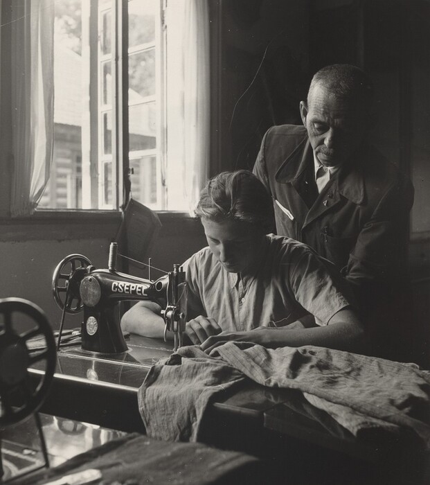 The image depicts two individuals from the waist up. The first person, a young woman, is seated at a sewing machine with a focused expression. She has fair skin and light hair pulled back. She wears a light-colored short-sleeved shirt. A man stands next to her, observing closely. He has short hair and a mustache, wearing a dark suit with a tie and a pin or badge on his lapel. The room has a large window on the left, providing natural light. Outside, a building and a tree are visible. Various fabric pieces are spread on the table.
