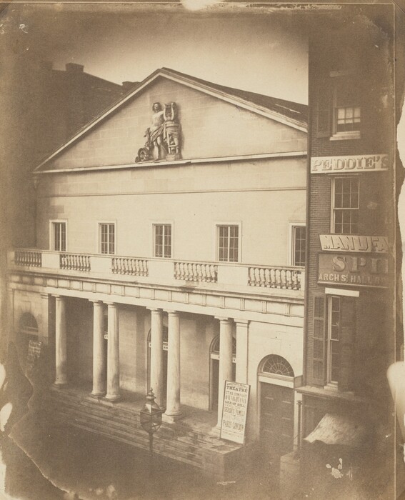 This is a photograph of a neoclassical building with columns and a pediment. The building features a façade with columns supporting a triangular pediment, adorned with a sculptural relief depicting a classical figure with a shield and a chariot. Below the pediment are rectangular windows, and beneath them is a balcony with a balustrade railing. A lamp post can be seen near the entrance, with steps leading up to several doorways. Signs indicate it is likely a public venue, possibly a theater, given the poster advertising performances. The photograph is in sepia tone, giving it a historic quality.