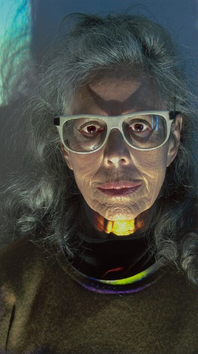 The image shows the upper half of a person's body, particularly their face. The person is upright and facing the camera, with identifiable facial features and visible wrinkles. They wear large, striped glasses and have silver-gray hair. The person is dressed in a brown sweater with a darker collar peeking through. Light and color are used effectively in the photograph, with reflections of blue, green, and purple on the clothing and neck. The background is plain blue.
