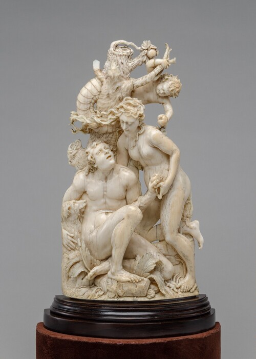 This sculpture depicts multiple people in a detailed scene. In the center, there are two people, one male and one female, both nude. They rest against the base of a tree with their hands together, holding an apple. The woman's long hair blows across her face, and the man looks up at her. To the left of the man is a dog, and below them are rocks, leaves, and shells. Above them, hanging from the top of the tree, is a child with the tail of a snake, holding another apple. The sculpture has a smooth texture and is an off-white color. It sits on a dark, round pedestal.