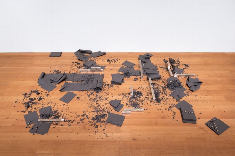 Wide strips of gray felt, lengths of square aluminum shafts, silver ball bearings, and felt cut up like confetti are scattered across a wood floor in this installation photograph. The aluminum lengths create verticals and horizontals. Some of the felt is cut into squares, which are stacked or splayed, or into longer lengths that crumple like cooked lasagna noodles. The shiny ball bearings are mostly in pairs or trios, carefully placed in rows or columns across the composition. From afar, the felt scraps resemble dirt, also strewn across the space.