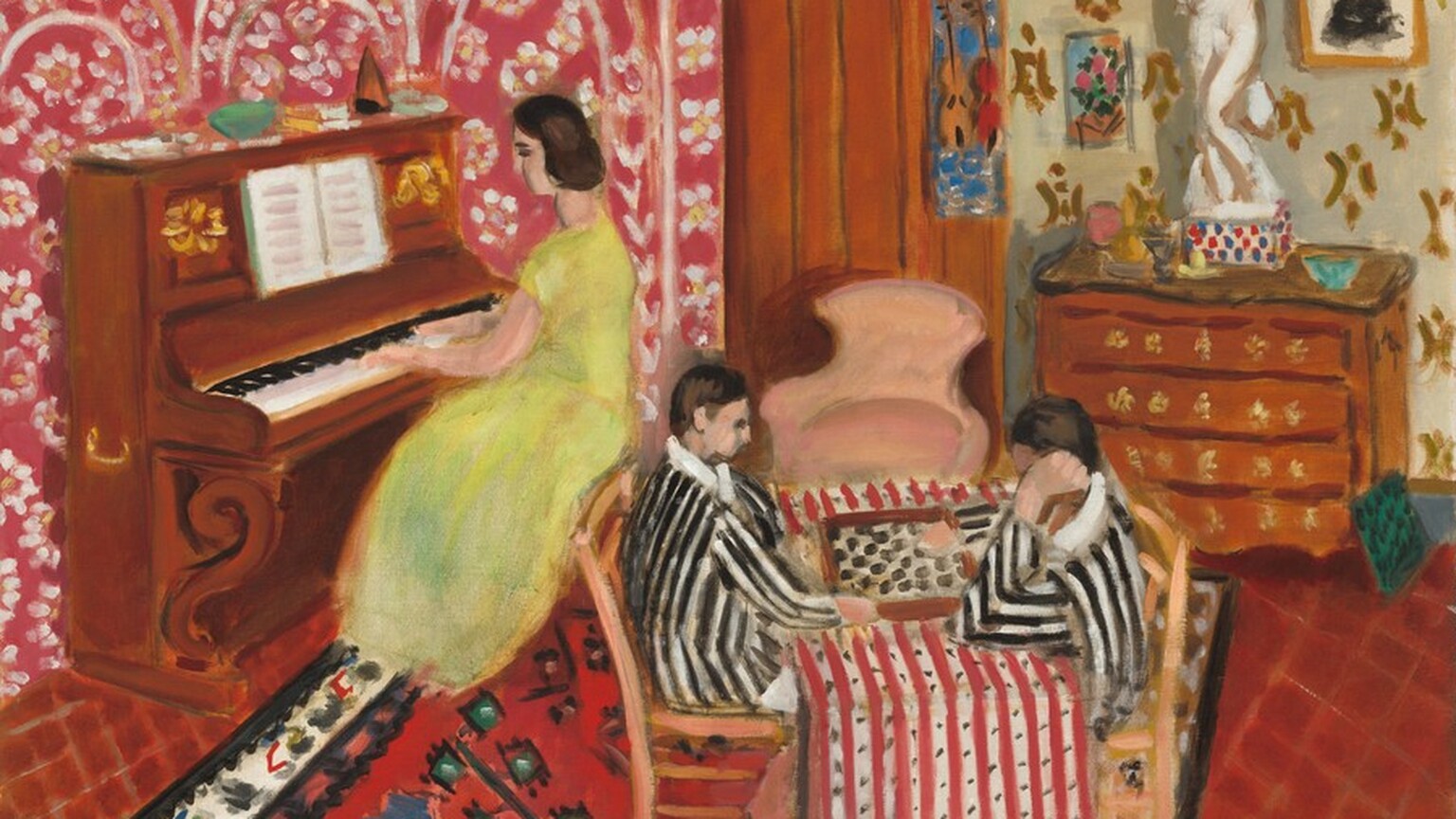 A woman plays an upright piano as two boys play checkers at a table in this horizontal painting. The scene is loosely painted with areas of flat color and pattern. The people have pale, peachy skin and dark brown hair. Their facial features are painted with dots or short strokes of black. The piano is against the wall to our left. The woman wears a short-sleeved, lemon-yellow dress with a long skirt. A music book is propped on the music stand, and loosely painted objects line the top of the instrument. The wall behind the piano is patterned with white and pale yellow flowers under and around arches against a cranberry-red background. The table where the boys play is in the middle of the room. The players sit in wooden chairs and wear black and white striped jackets with white collars. The boy to our right looks at the board while resting his left cheek, closer to us, in his hand. The other boy looks on with his arms on the table in front of him. The checkerboard sits on a red and white striped cloth, which has lines of black dots in the white strips. Beneath the table is a floral rug with blue and black flowers against a red background. The border is painted loosely with leafy shapes against bright white. Beneath the area rug the floor or another carpet is garnet red with a diamond-shaped grid painted in orange. A petal-pink, upholstered chair sits in front of a tall wardrobe in the back corner of the room. A picture with two violins against a blue background hangs on the side of the furniture to our right. A chest of drawers next to the wardrobe holds a few bowls or objects and a white statue, about half human height. The statue is of a person standing with one knee bent in front of the other, one elbow tucked by the side with that hand held to the chest, and the other bent elbow raised overhead. Pictures hang on the wall around the statue, and the wallpaper there is ivory white with a dark yellow pattern. The artist signed the work in the lower right corner, “Henri Matisse.”