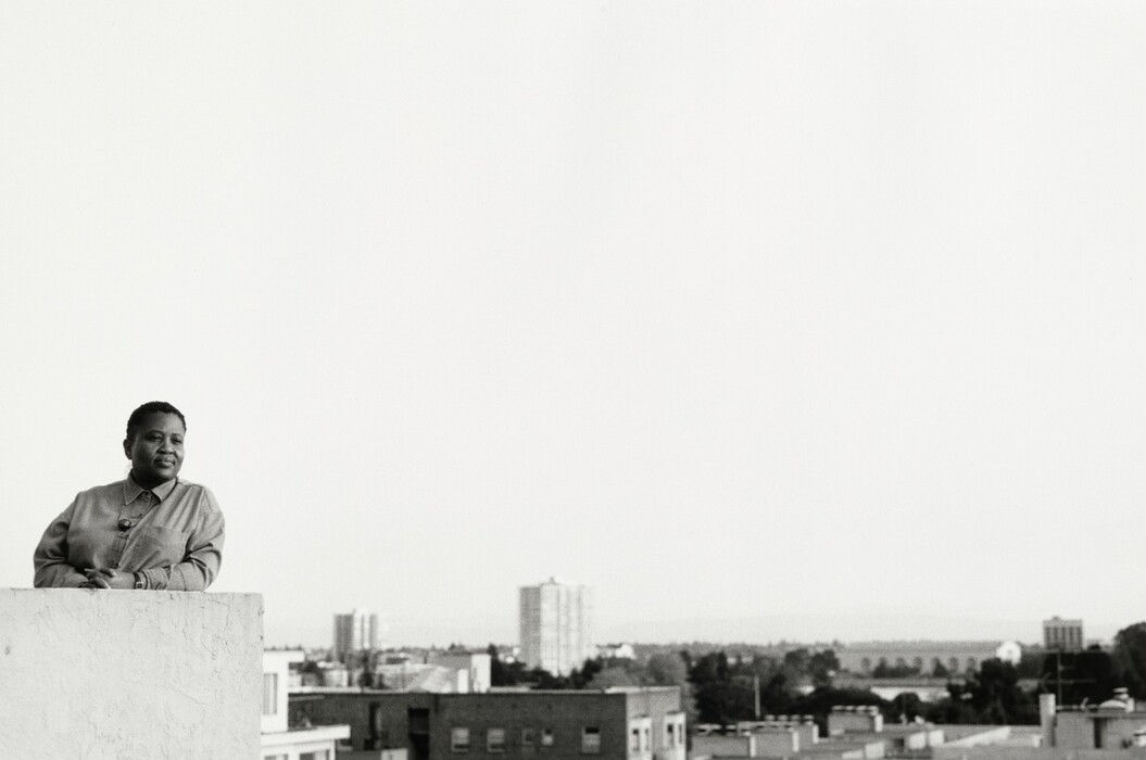 The image shows a person from the hips up, leaning on a ledge or parapet. The person has short hair and wears a collared, long-sleeved shirt. They appear calm or contemplative, with hands resting on the ledge. A watch is visible on their left wrist. The background features an urban landscape with buildings in the distance, against a sky. The image is in black and white.