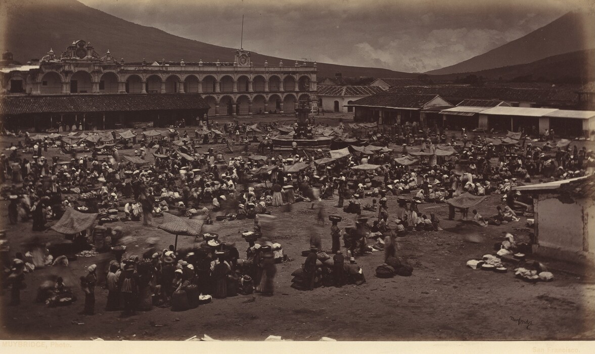 The image shows a busy market scene from an elevated point of view. The horizon is hidden by mountain slopes, enclosing the market area. A grand colonial building with arches and a decorative facade is visible in the background. The photograph is fine and detailed, with a sepia color palette. Many people are present in the market, some stationary and others in motion. More individuals are seen under canopies in the distance.