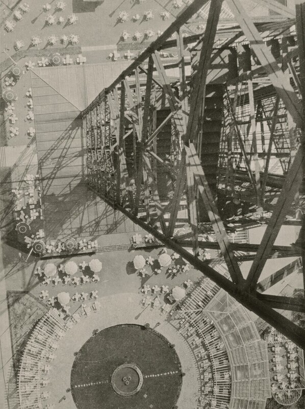 From very high up, we look down along the height of a metal radio tower at a plaza on the ground, which is dotted with umbrellas, tables, and chairs in this vertical black and white photograph. The side of the tower fills almost the height of the right edge of this image. Crisscrossing girders form diamond shapes down the sides, enclosing stairways and other structures. In the plaza below, the tables and chairs create cross and asterisk shapes around the disks of the umbrellas, seen from above. Along the bottom of the photograph, and cut off by that edge, the tables and a skeletal awning creates a ring around a circular area, presumably grass.