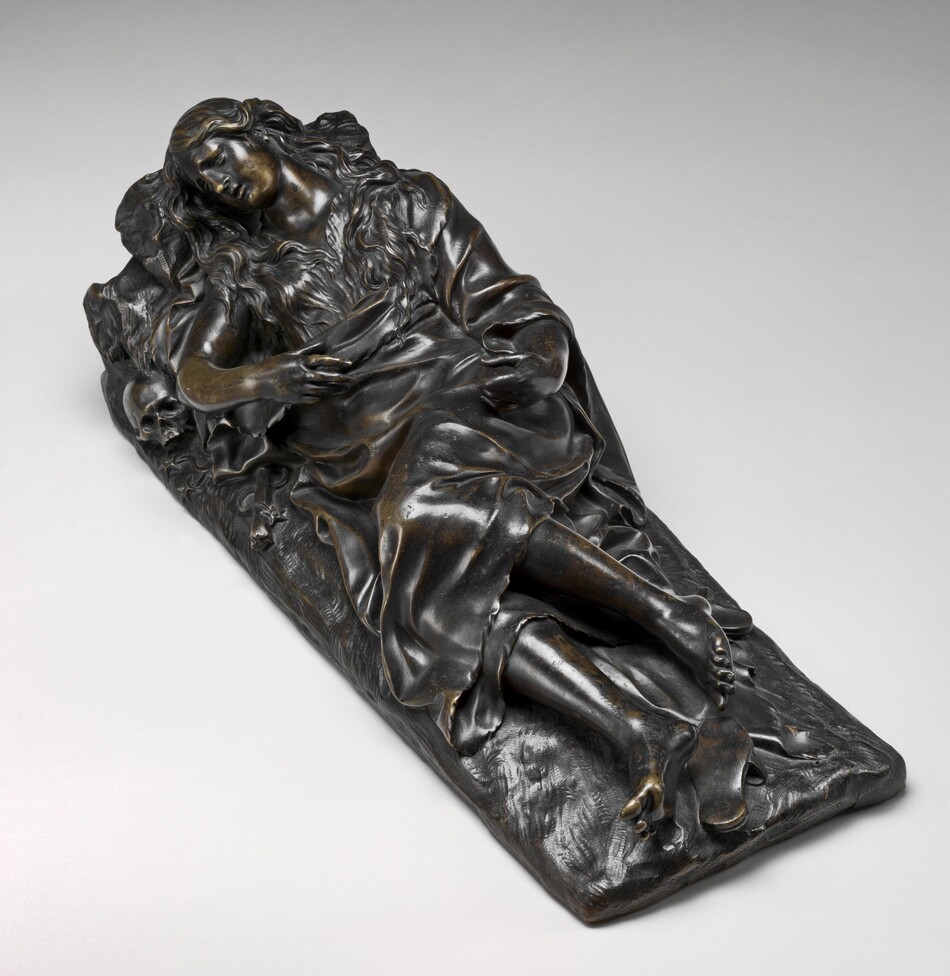 The sculpture depicts a reclining female figure with long, wavy hair and ornate features. She is draped in flowing robes that cascade over a rocky surface beneath her. Her head rests upon a cushioned surface and is tilted to the side in a relaxed posture, and her arms are crossed over her waist. Adjacent to her left elbow, a skull is subtly integrated into the composition. The sculpture is made of bronze with a dark patina. The clothes are shiny, emphasizing their folds. The piece is intricately detailed, with realistic proportions and features.