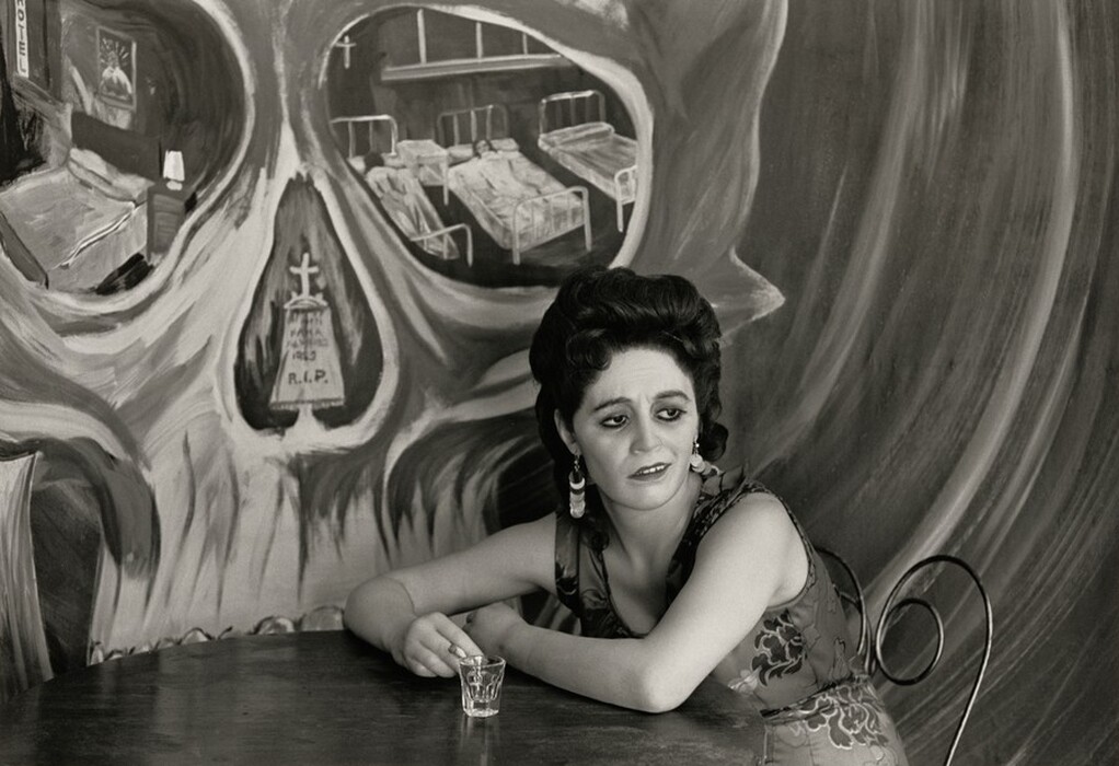 The image shows a woman seated at a table, captured from the waist up. She leans slightly forward with one arm rested on the table while holding a shot glass with the other hand. She has prominent eyebrows and deeply set eyes, with dark hair styled in a voluminous updo. She wears patterned clothing, possibly a sleeveless dress, along with large earrings. The background features an elaborate mural depicting a skull with scenes of beds and a tombstone with "R.I.P." Inside the skull's eye sockets.