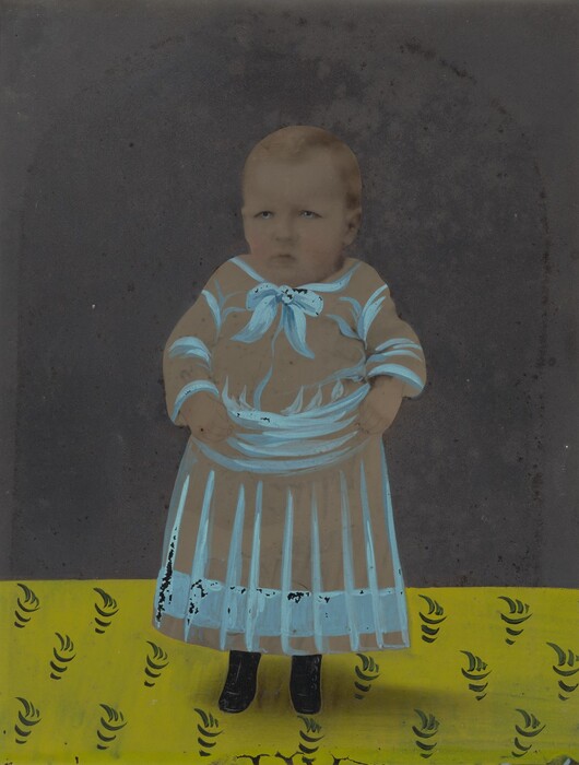 The image shows a young child standing upright. The child has a serious expression with short, light brown hair. They are wearing a long beige dress with blue accents, including a large blue bow at the neck and decorative blue patterns throughout. The child is standing on a yellow surface with black swirl patterns and the background is plain dark grey.