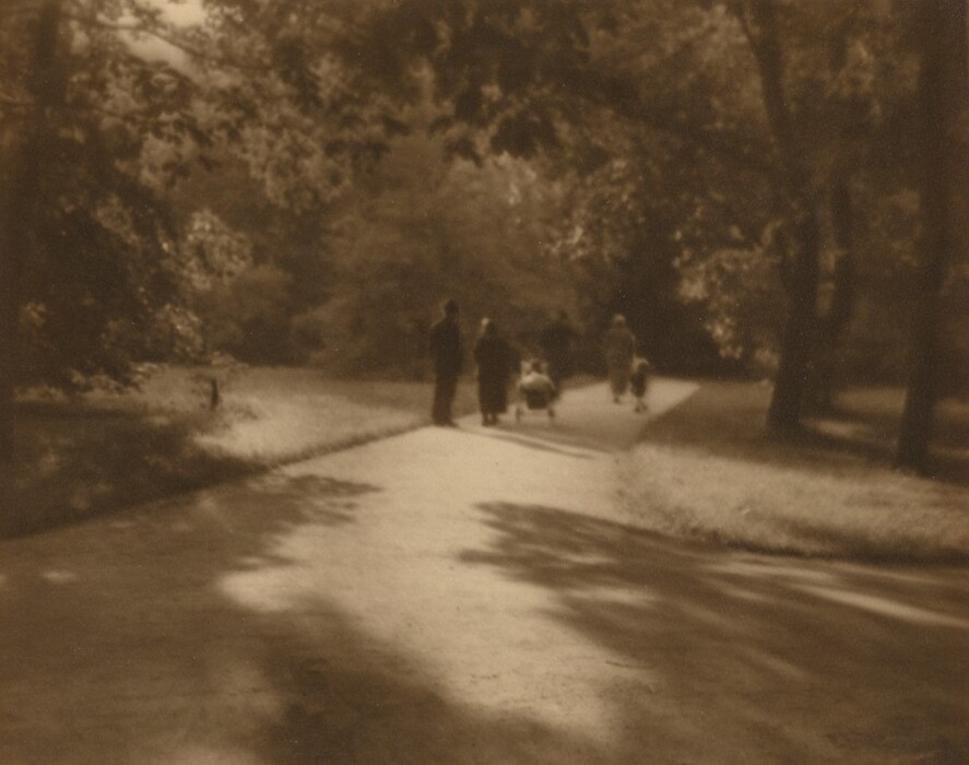 This blurry, sepia-toned photograph shows a view of a tree-lined path in a park. The photograph is taken from a low angle, looking down the path, which extends into the distance. Closer to us, the path is empty, but further along, there are people visible, including one person pushing a stroller. The people are blurred and indistinct. The path is bordered by grass and trees, with branches forming a canopy overhead. Light filters through the leaves, creating shadows.