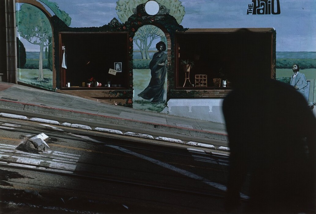 This is a photograph of an urban street scene with a large mural. The image captures a street that slopes downhill, with parts of the sidewalk and road visible, including tracks for a tram. On a building wall in the background, there is an intricate mural showing figures, trees, and a scenic landscape painted on it. The mural includes a person in a robe standing by an archway, surrounded by greenery and trees, creating an illusion of depth and altering the perception of space. The right side of the image is a dark silhouette, contrasting with the brightly lit mural and street surface. The lettering "The Patio" is visible on the upper right side of the mural, indicating possible significance to the mural's location. Elements like a window with plants and pots are integrated into the mural.