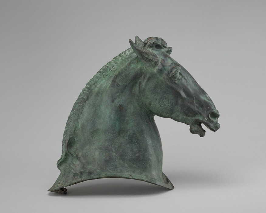 The sculpture is of a horse's head. It captures details such as the ears, eyes, nostrils, and a slightly open mouth. The sculpture is made of dark metal, likely bronze, with a green patina. It has a textured and aged appearance. The sculpture is presented in profile view, emphasizing the contours and musculature of the horse's neck and head, as well as the intricately carved mane along the top. The naturalistic portrayal of the horse's features gives the sculpture a lifelike and dynamic presence.