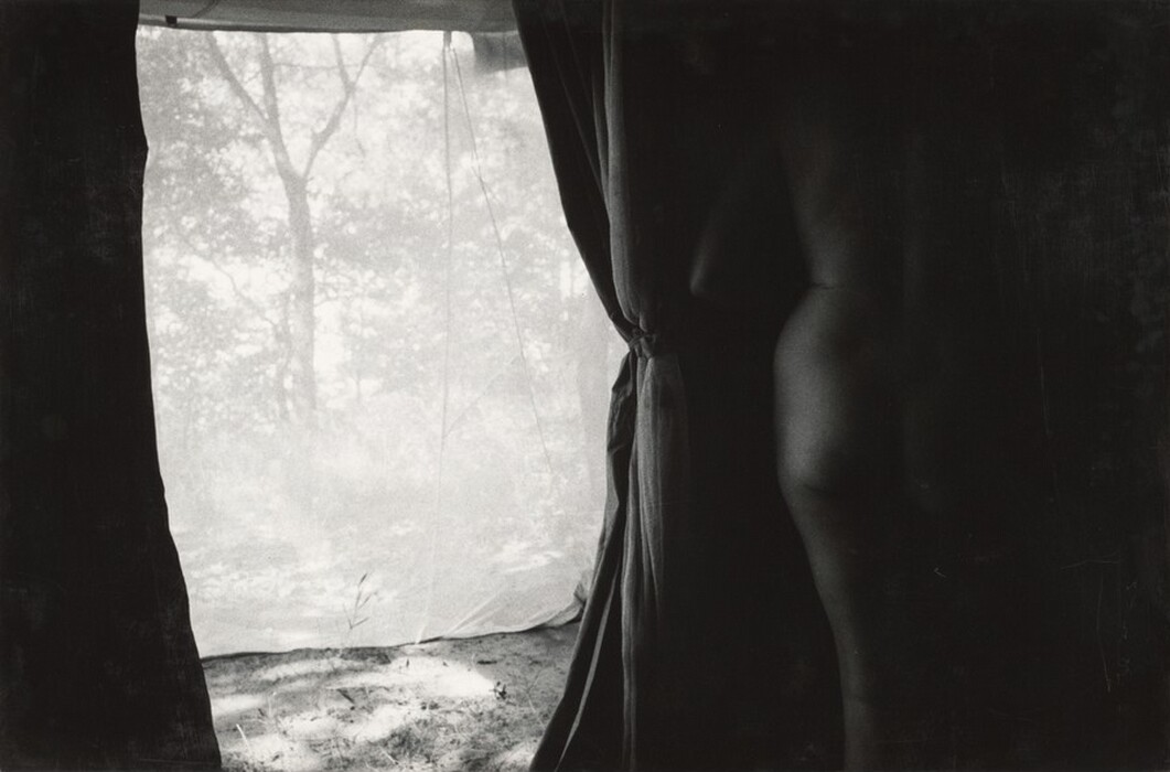 The image shows a partial view of a person's body focusing on their lower back, hip, and upper leg area. The person is standing to the right of the frame with their back facing towards the viewer. There are no visible facial features or hair, as only the lower portion of the body is captured in the shadowed area. The person appears to be nude and is standing next to a curtain or fabric that is tied to the right side, adding to the shadow effect. The background consists of a translucent material, possibly another sheet or tent-like fabric, through which silhouettes of trees and foliage can be seen against the light. The ground in the immediate foreground appears to be uneven and illuminated, suggesting some outdoor setting behind the translucent fabric.